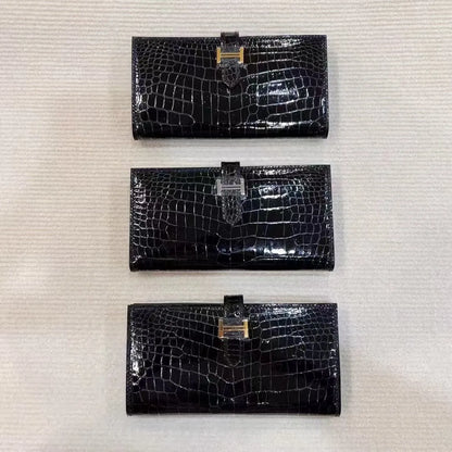 Three black crocodile-embossed leather wallet clutches are lined up vertically on a textured off-white background, each featuring a gold-tone H-shaped clasp.