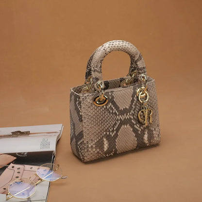 A structured snakeskin handbag with a top handle and gold-tone hardware, styled on a warm tan background with an open magazine and round glasses nearby.