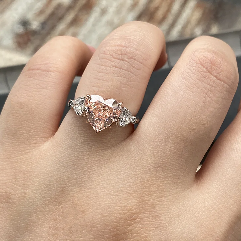 A close-up of a hand wearing a silver ring with a large heart-shaped pink gemstone and smaller clear stones on either side.