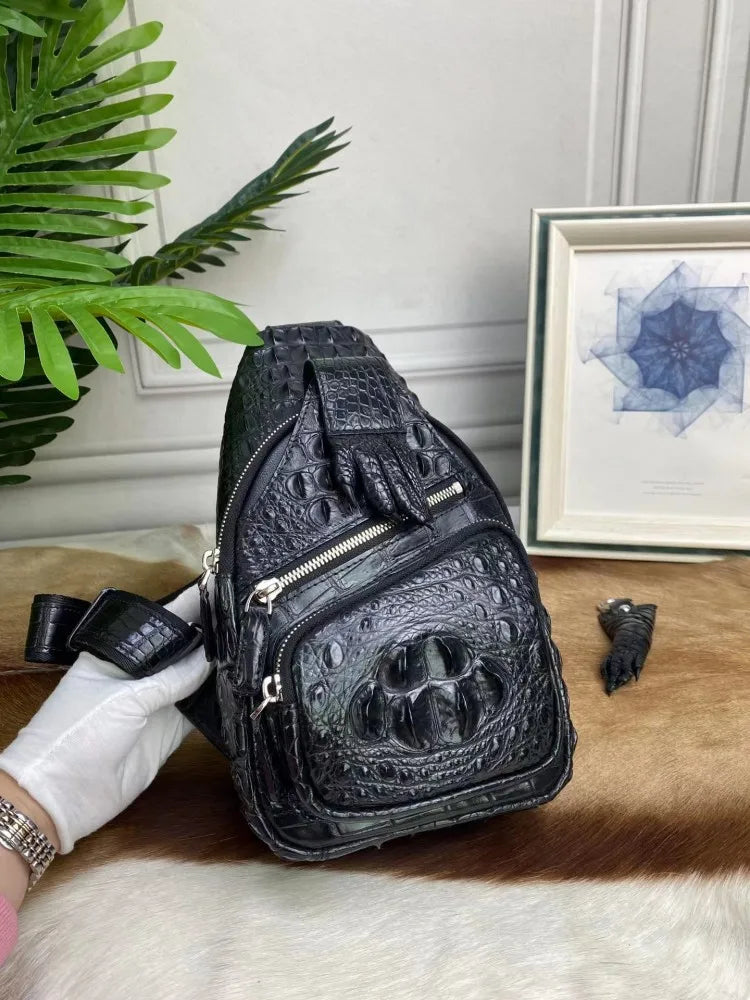A black crocodile-embossed leather crossbody bag with a prominent crocodile head motif on the front pocket, held by a hand wearing a white glove. The bag is displayed on a brown and white faux fur surface with a green plant and a framed picture in the background.