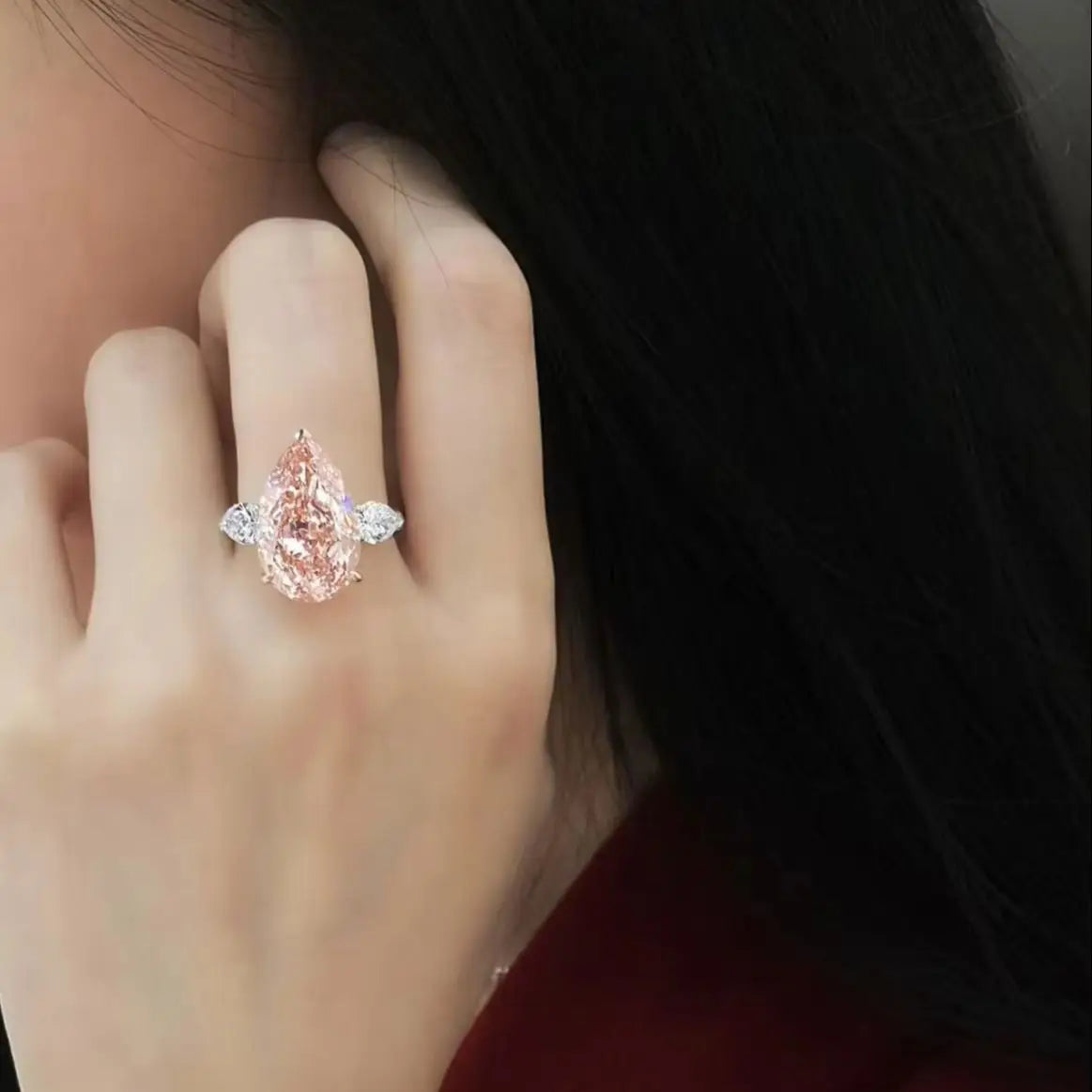 A close-up of a hand wearing a silver ring with a large pear-cut pink gemstone and smaller side stones, set against dark hair and a red garment.