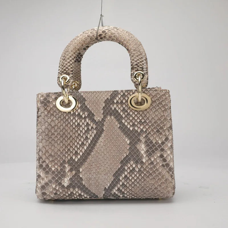 A structured handbag with a snakeskin pattern in beige and gray, featuring a rounded top handle and gold-tone hardware, photographed against a neutral light gray background.
