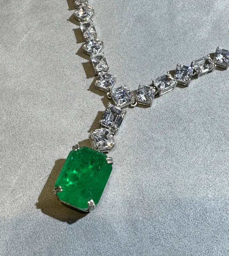 A silver-toned Y-shaped necklace featuring a large rectangular emerald-cut green gemstone pendant and a line of clear square-cut stones along the chain, displayed on a textured gray surface.