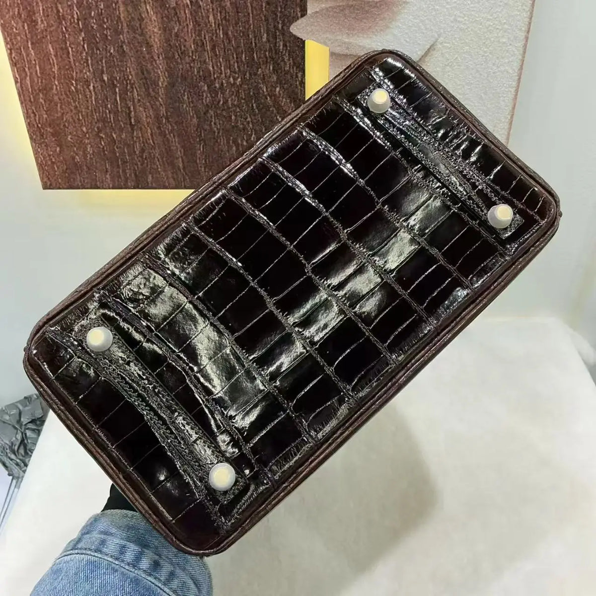 A dark brown crocodile-embossed leather handbag is held from below, showing its glossy, textured bottom with four white circular studs. A person's denim-clad hand is visible holding the bag, set against a background of a white surface and a wooden panel.