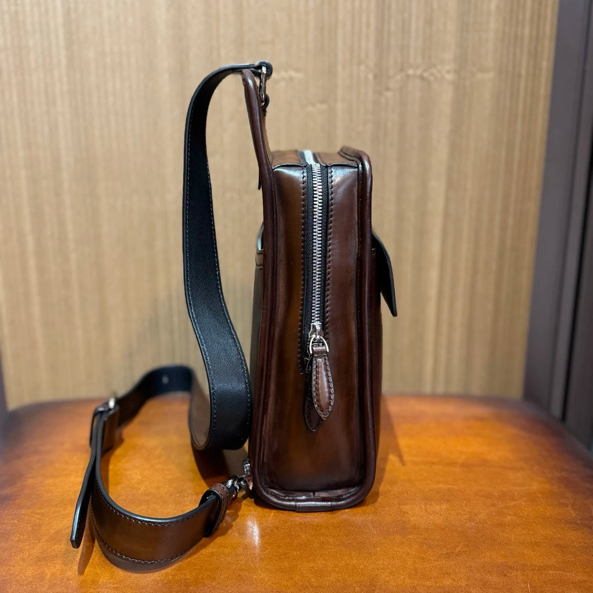 A dark brown leather crossbody bag with a black shoulder strap, standing upright on a wooden table against a wood-paneled background.