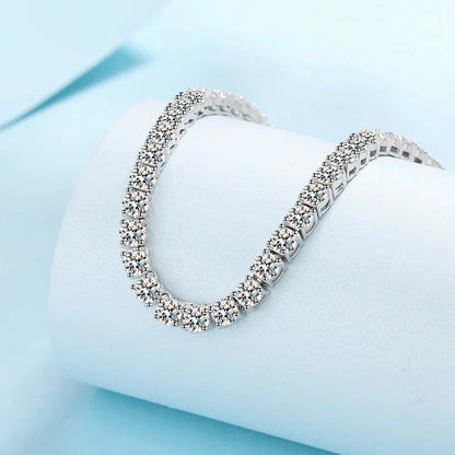 A silver tennis bracelet paved with clear stones, displayed on a white cylindrical jewelry stand against a light blue background.