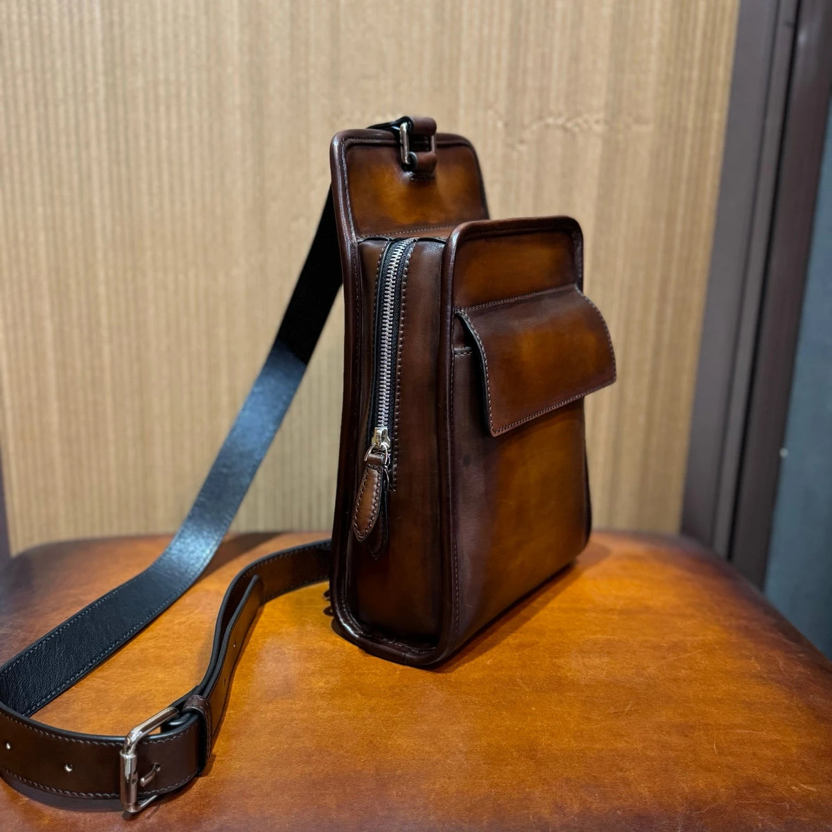 A brown leather crossbody bag with a black adjustable strap, standing on a wooden surface against a wood-paneled background.