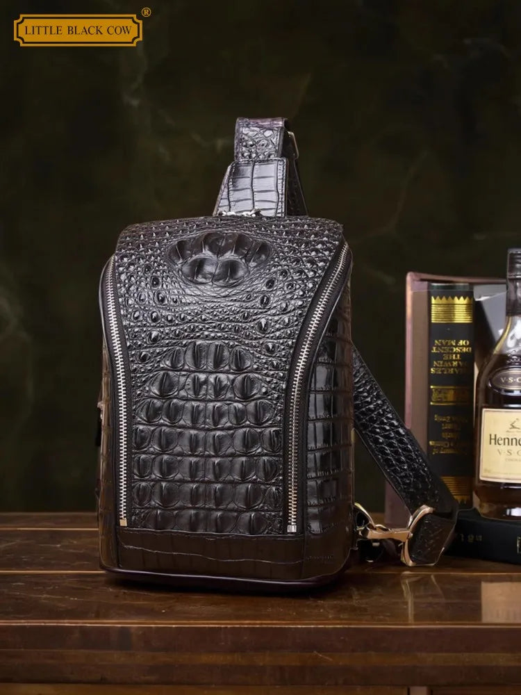 A dark brown crocodile-embossed leather crossbody bag with a top handle and shoulder strap, displayed on a wooden table next to books and a bottle, with a 'Little Black Cow' logo in the top left corner.