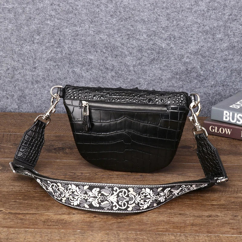 Black faux-crocodile leather half-moon shoulder bag with a wide, embellished silver and black floral strap, resting on a wooden surface against a gray felt background.