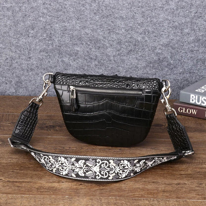 Black faux-crocodile leather half-moon shoulder bag with a wide, embellished silver and black floral strap, resting on a wooden surface against a gray felt background.