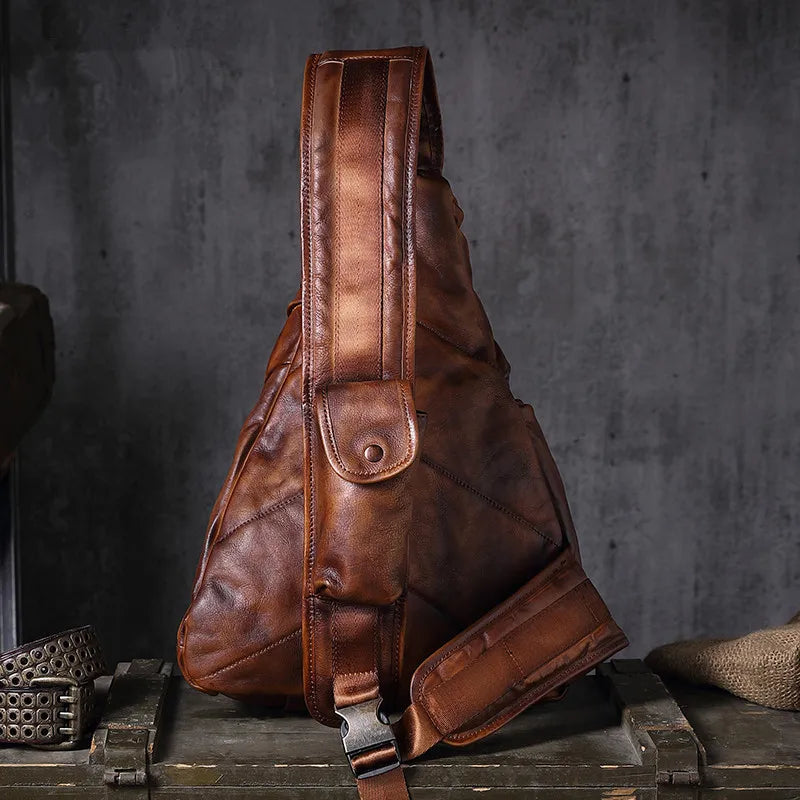 A brown distressed leather sling bag with a single shoulder strap, displayed on a rustic wooden crate against a dark gray concrete wall.