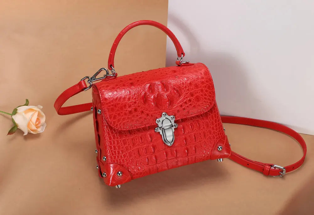A bright red crocodile-embossed leather handbag with a top handle, shoulder strap, and silver-tone hardware, styled on a beige background with a small rose prop.
